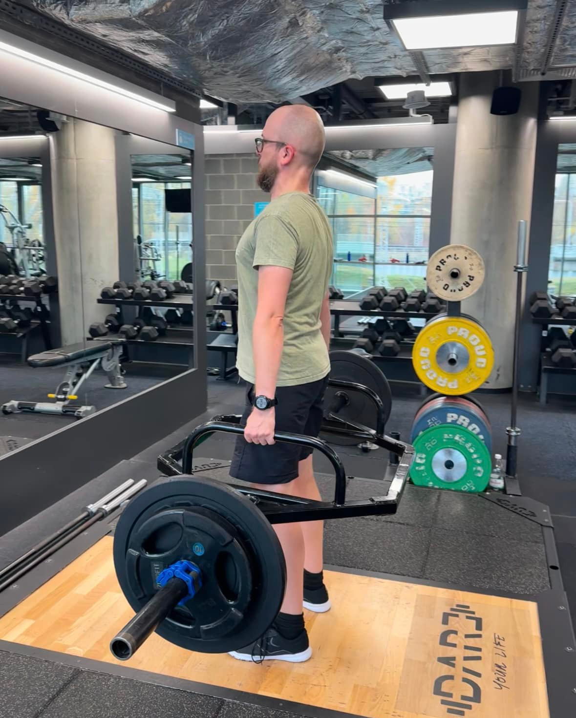 Mężczyzna w okularach podnosi ciężar na specjalnym gryfie w siłowni. Widoczne obciążenia, lustro i sprzęt do ćwiczeń. Trening siłowy.