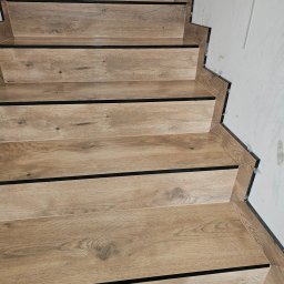 REMMARO - Wykończone schody z płytek imitujących drewno, z czarnymi listwami, przy białej ścianie w trakcie wykończenia. Detal wykończenia schodów.