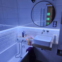 REMMARO - Nowoczesna łazienka z białymi płytkami, podświetleniem LED i okrągłym lustrem. Umywalka nablatowa na drewnianym blacie. Widoczne akcesoria łazienkowe i oświetlenie punktowe.