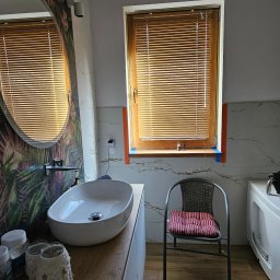 REMMARO - Nowoczesna łazienka z owalnym lustrem, drewnianymi żaluzjami i marmurowymi płytkami. Umywalka nablatowa i dekoracje w stylu boho dodają wnętrzu unikalnego charakteru.