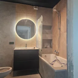 Nowoczesna łazienka z okrągłym lustrem podświetlanym LED, szarą glazurą i wanną. Elementy zabezpieczone folią ochronną. Widoczne bidet i czarne szafki.