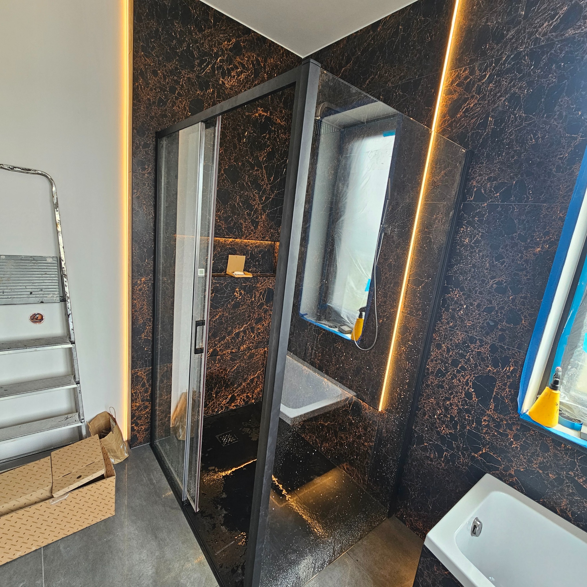 Wykończona łazienka z czarnymi płytkami imitującymi marmur, kabiną prysznicową z czarną ramą i podświetleniem LED. Widoczna drabina i zabezpieczona folią wanna.