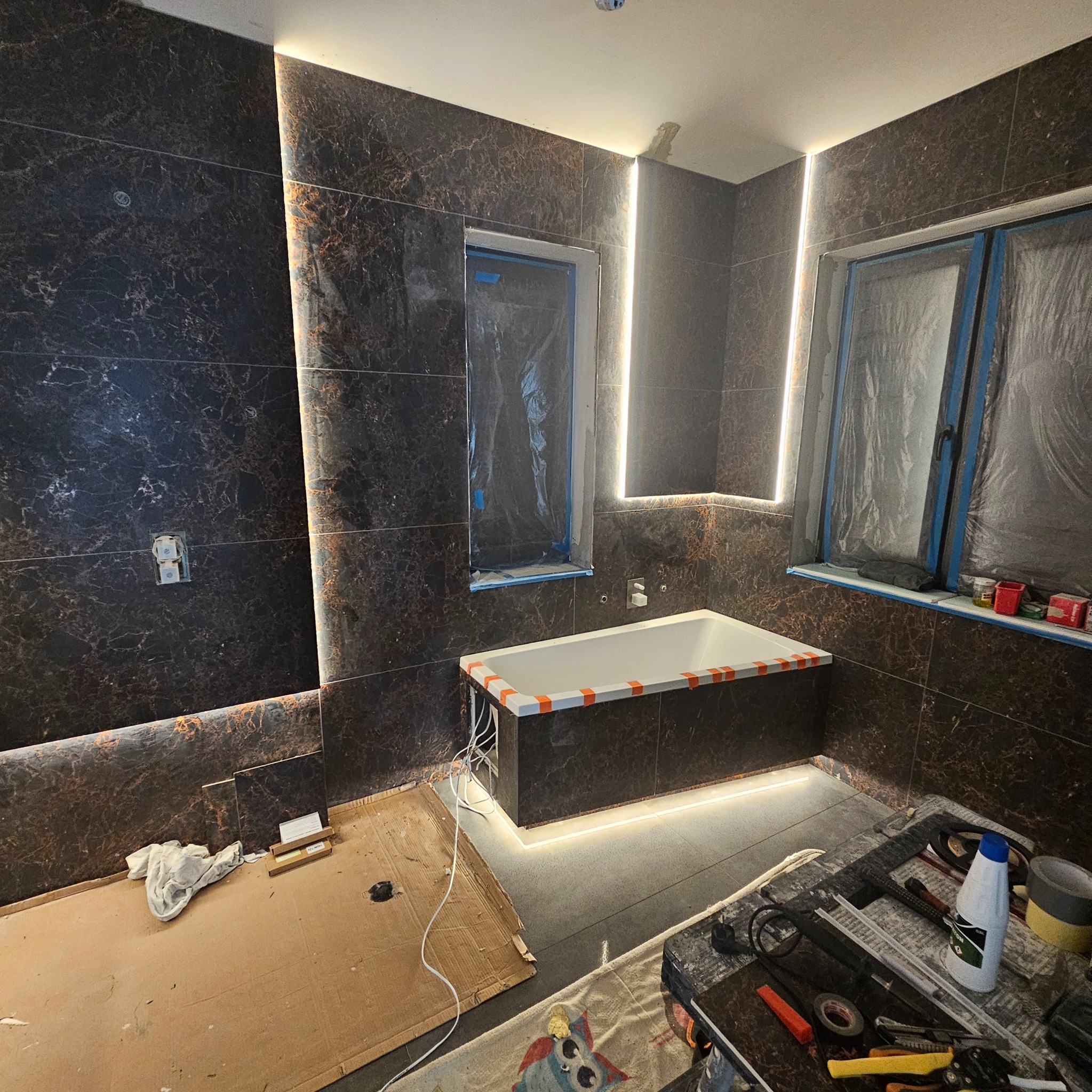 Montaż płytek imitujących marmur w łazience z podświetleniem LED. Okna zabezpieczone folią. Widoczne narzędzia glazurnicze i fragment wanny.