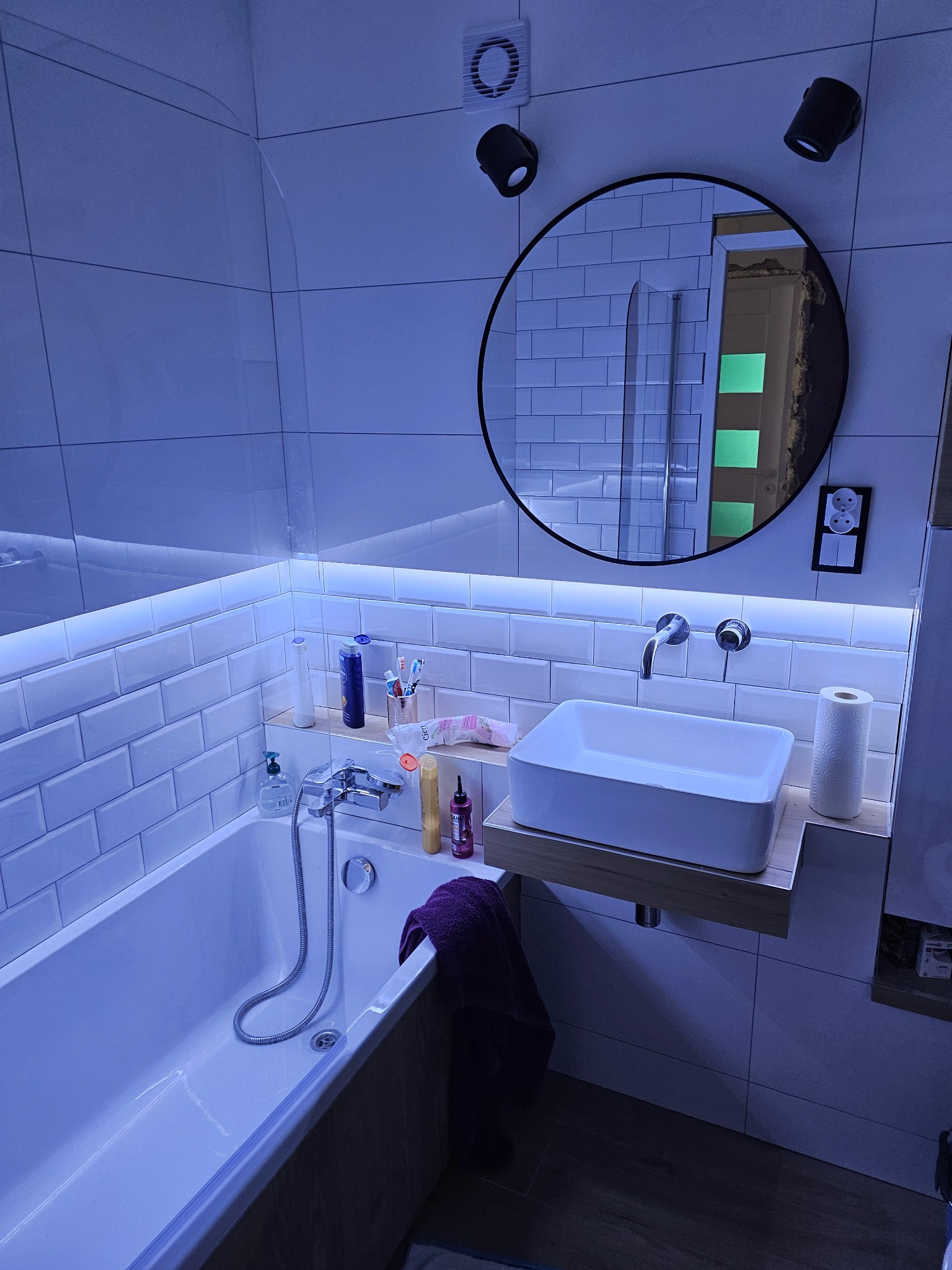 Nowoczesna łazienka z białymi płytkami, podświetleniem LED i okrągłym lustrem. Umywalka nablatowa na drewnianym blacie. Widoczne akcesoria łazienkowe i oświetlenie punktowe.