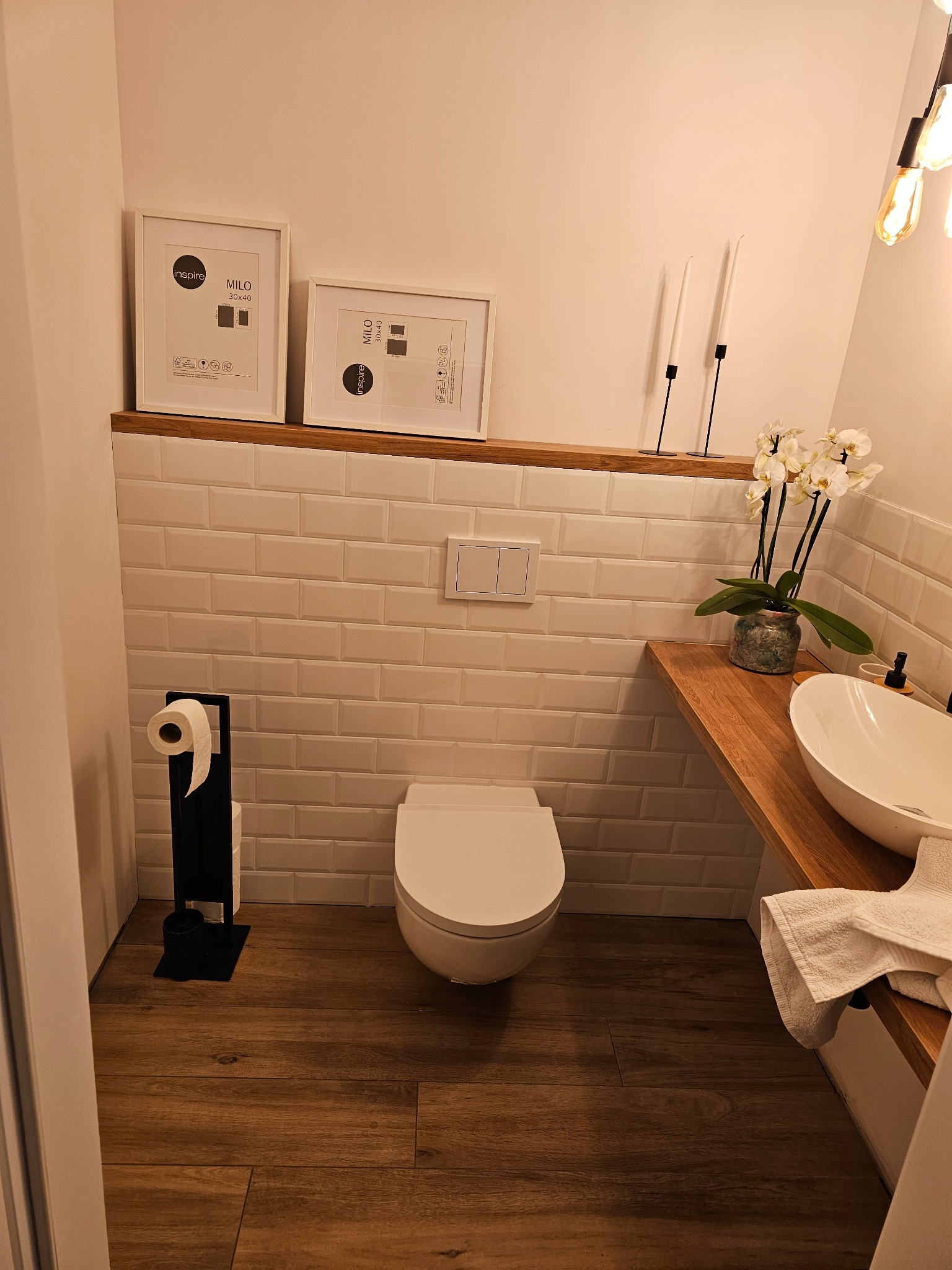 Nowoczesna łazienka z drewnianą podłogą i blatem, białą armaturą, płytkami typu metro oraz dekoracjami: ramki, świece i orchidea.