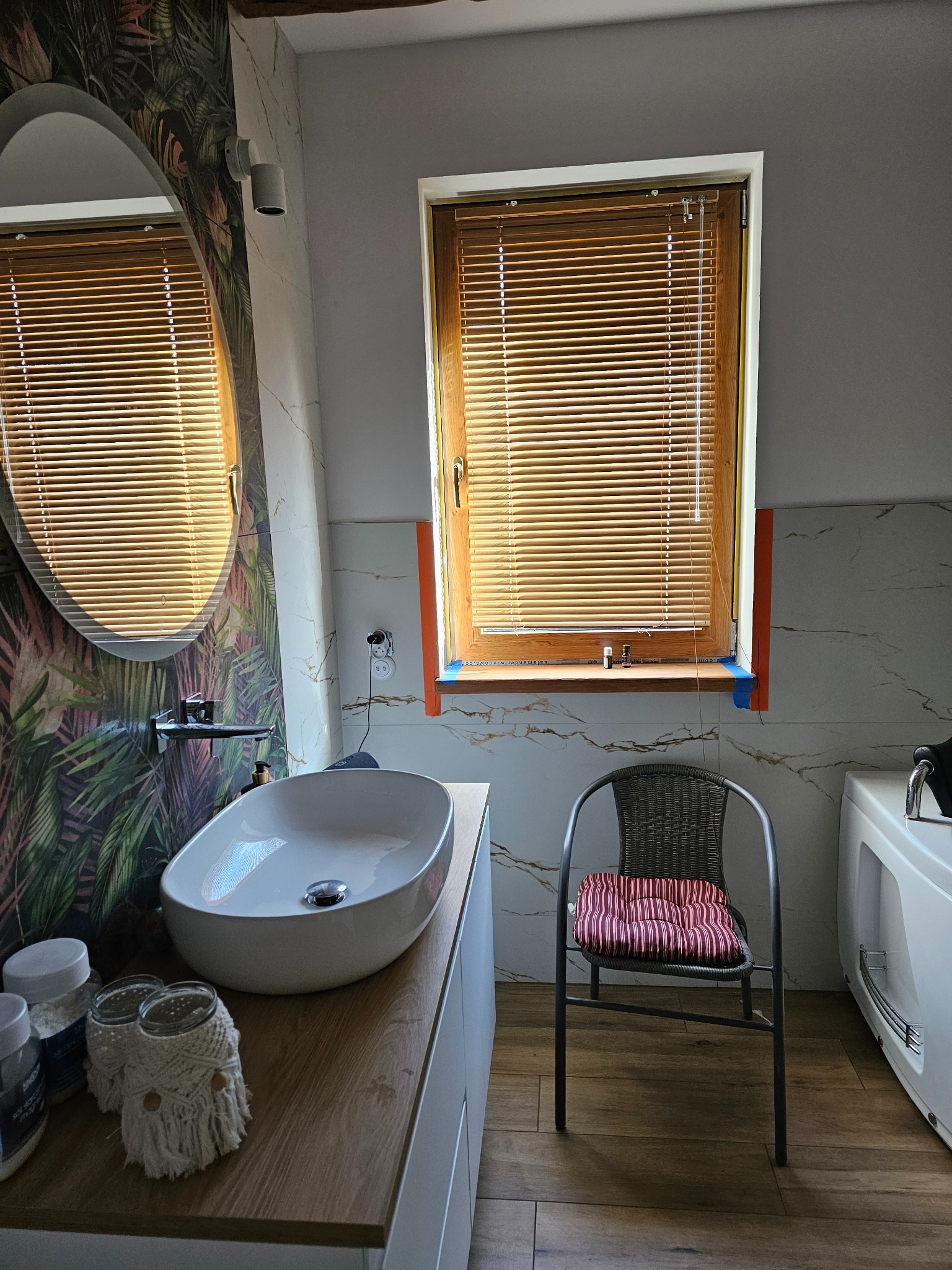 Nowoczesna łazienka z owalnym lustrem, drewnianymi żaluzjami i marmurowymi płytkami. Umywalka nablatowa i dekoracje w stylu boho dodają wnętrzu unikalnego charakteru.