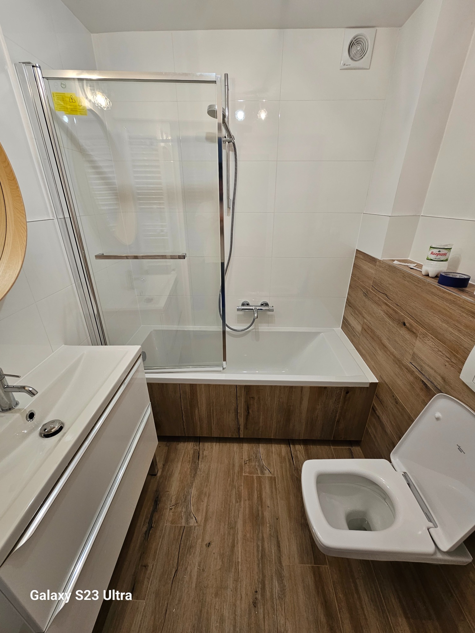 Nowoczesna łazienka po remoncie: białe płytki, drewnopodobne panele, kabina prysznicowa, umywalka z szafką i podwieszana toaleta. Widoczny wentylator sufitowy.