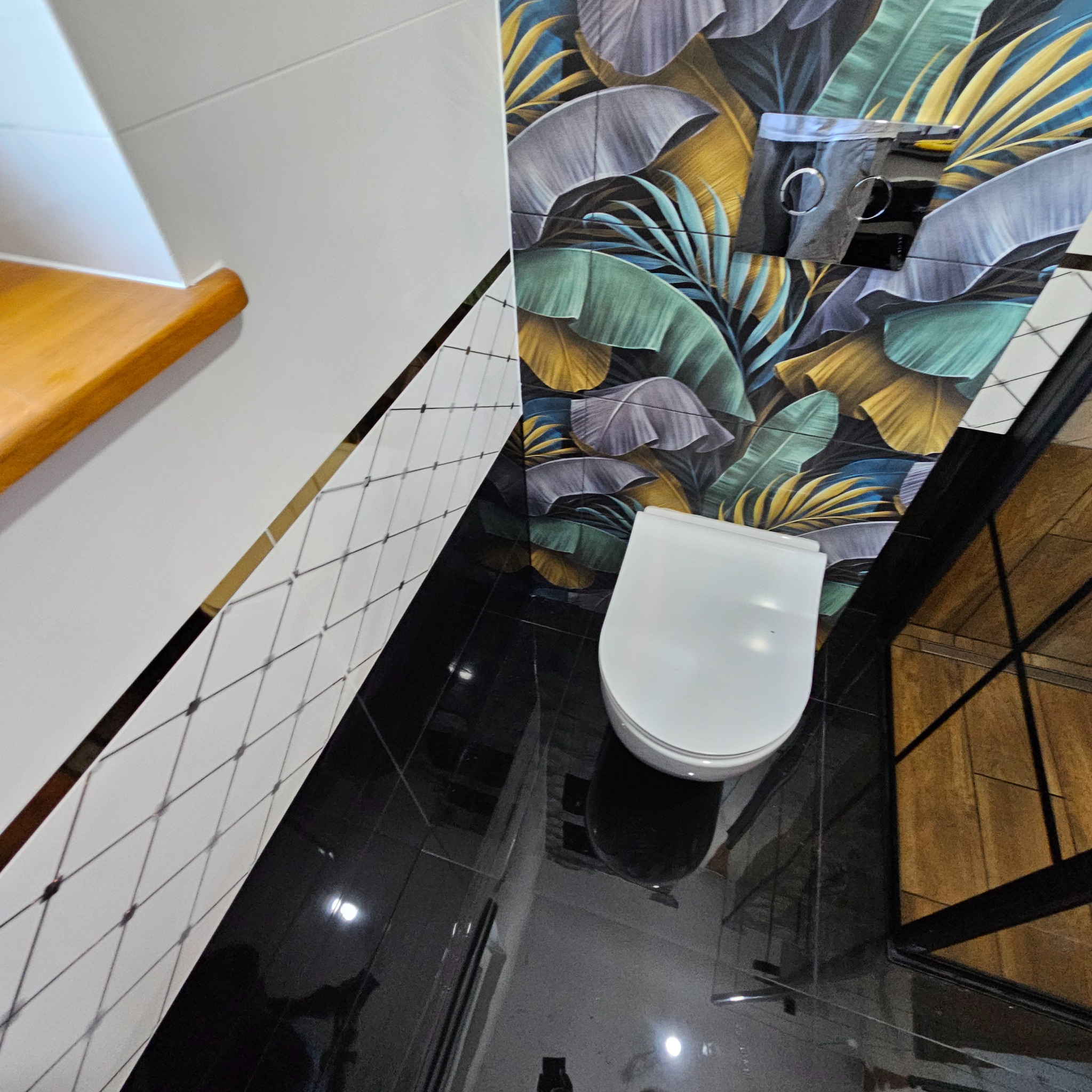 Nowoczesna łazienka z czarną glazurą, toaleta podwieszana i ściana w tropikalne liście. Elementy drewniane. Ujęcie z góry.