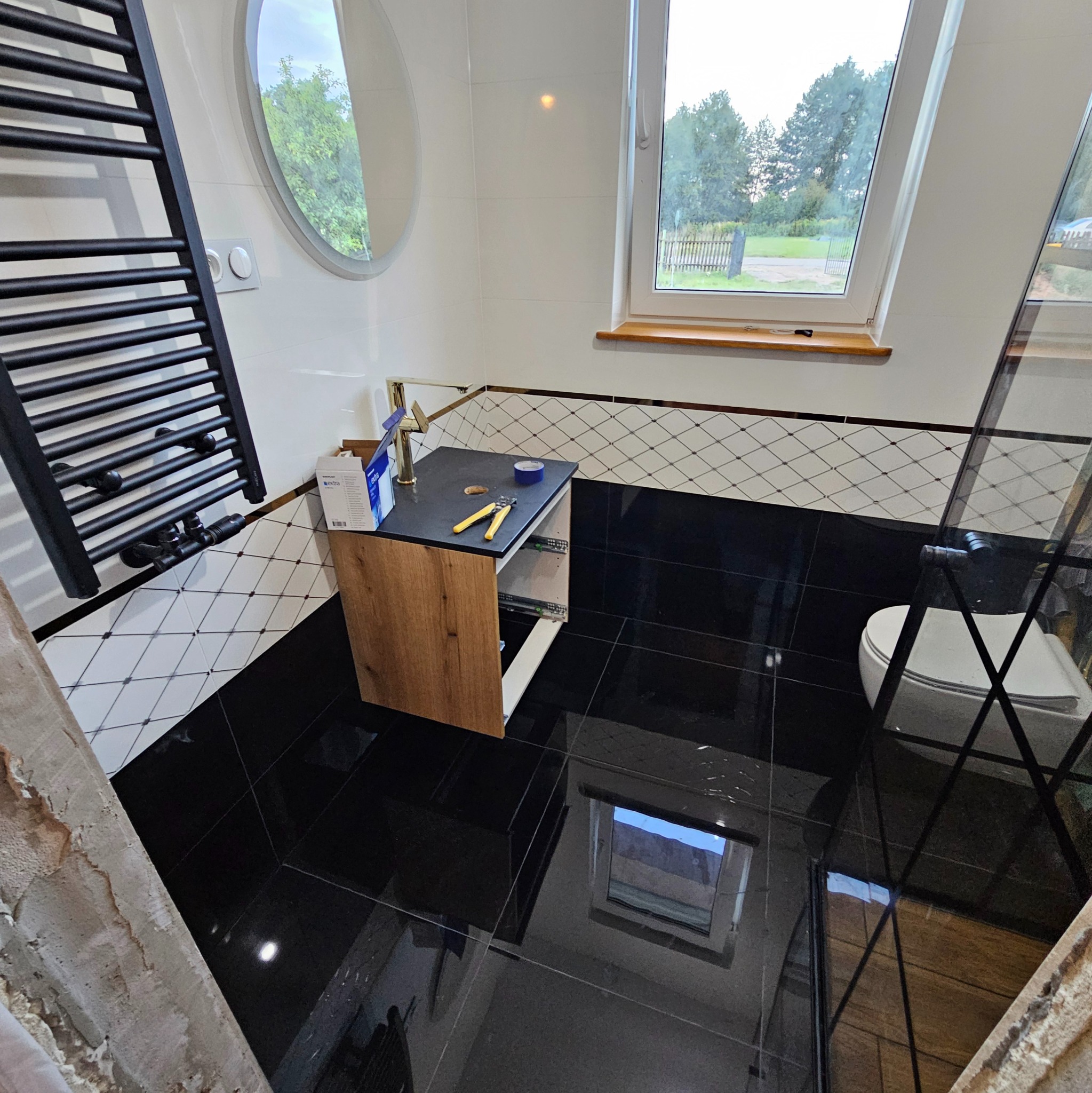 Częściowo wykończona łazienka: czarne płytki podłogowe, białe ściany z dekoracyjnym pasem, drewniana szafka pod umywalkę, grzejnik drabinkowy i okrągłe lustro.