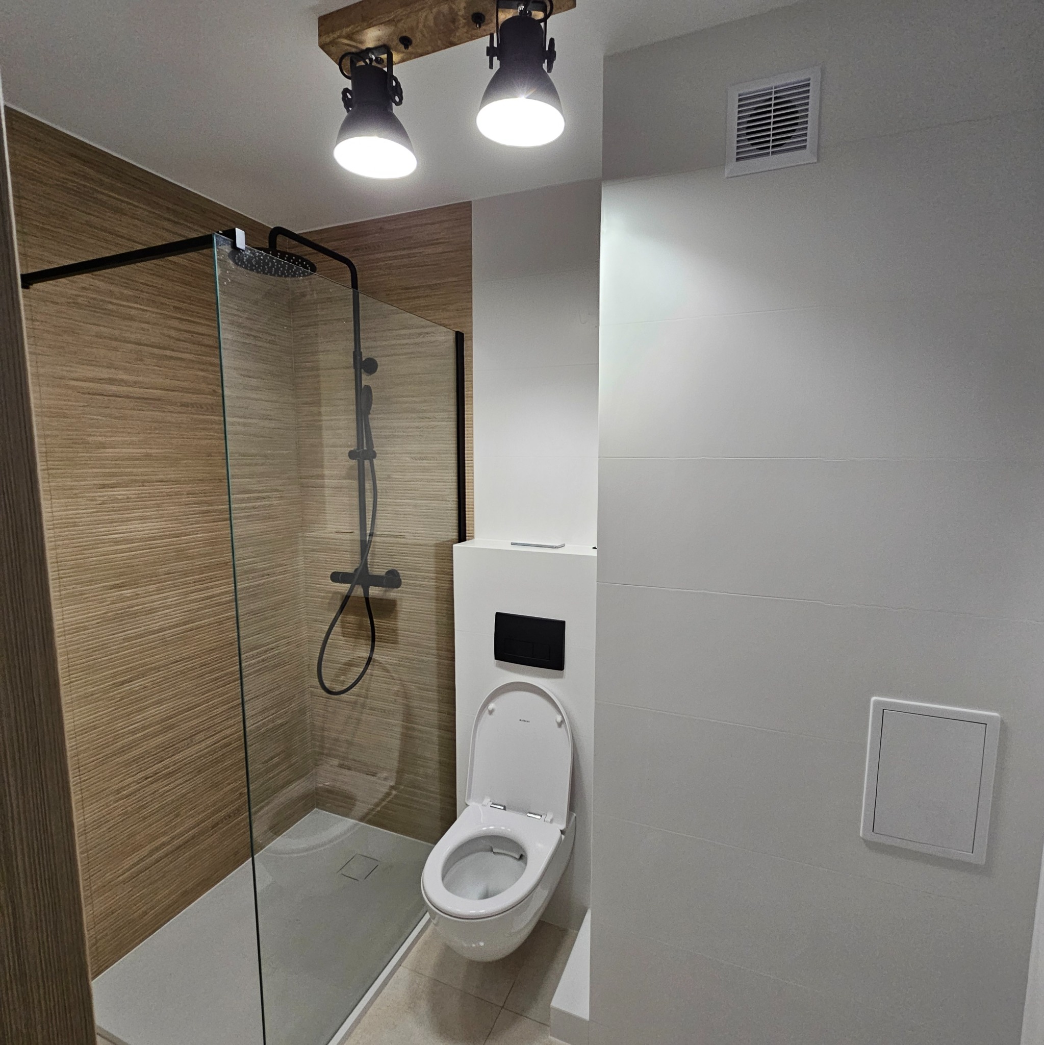 Nowoczesna łazienka po remoncie: kabina prysznicowa typu walk-in z czarną armaturą, podwieszana toaleta i oświetlenie punktowe.