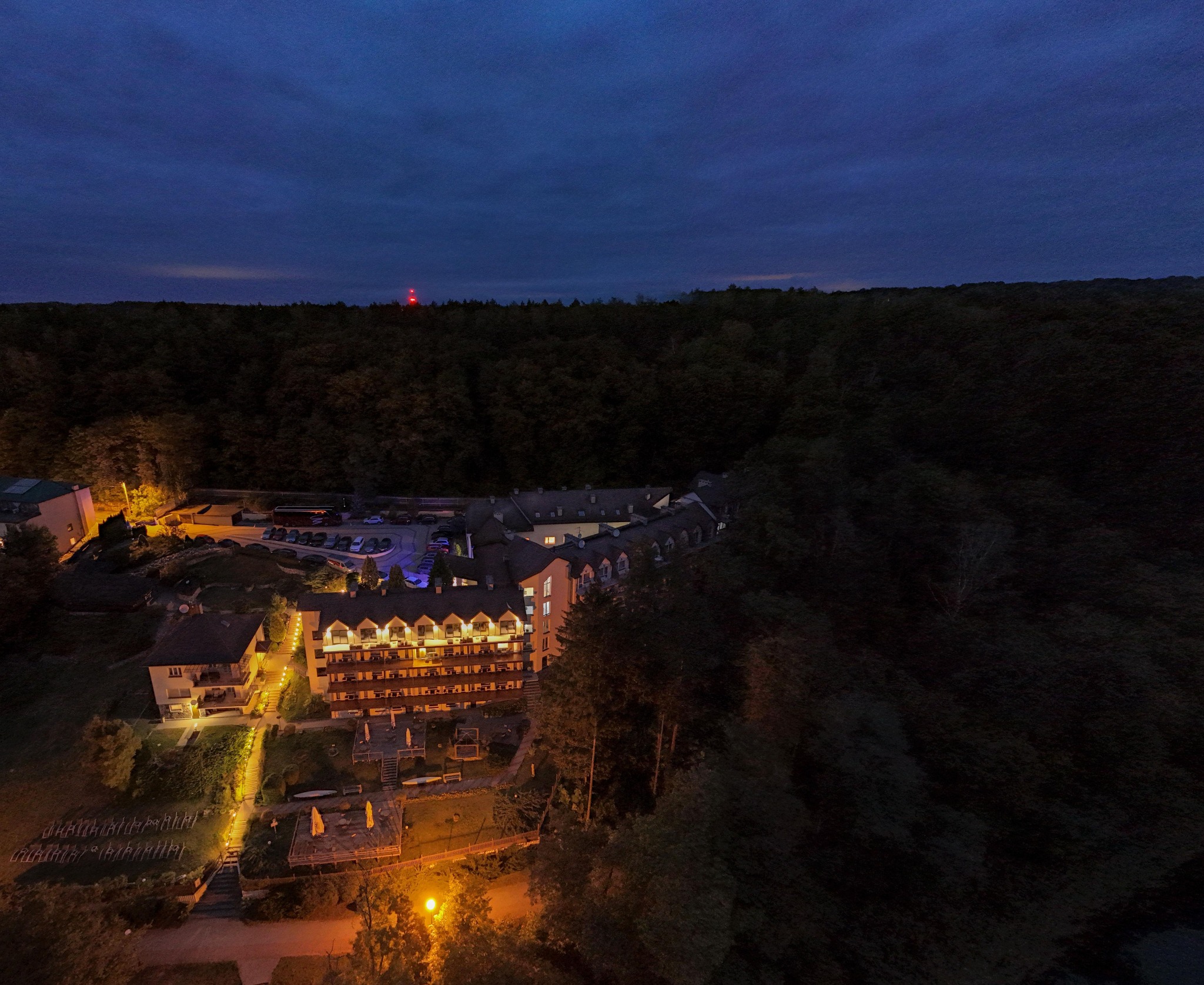 Nocny widok z lotu ptaka na oświetlony kompleks hotelowy z parkingiem i zielonym terenem rekreacyjnym otoczony ciemnym lasem, na horyzoncie widoczna czerwona lampa ostrzegawcza.