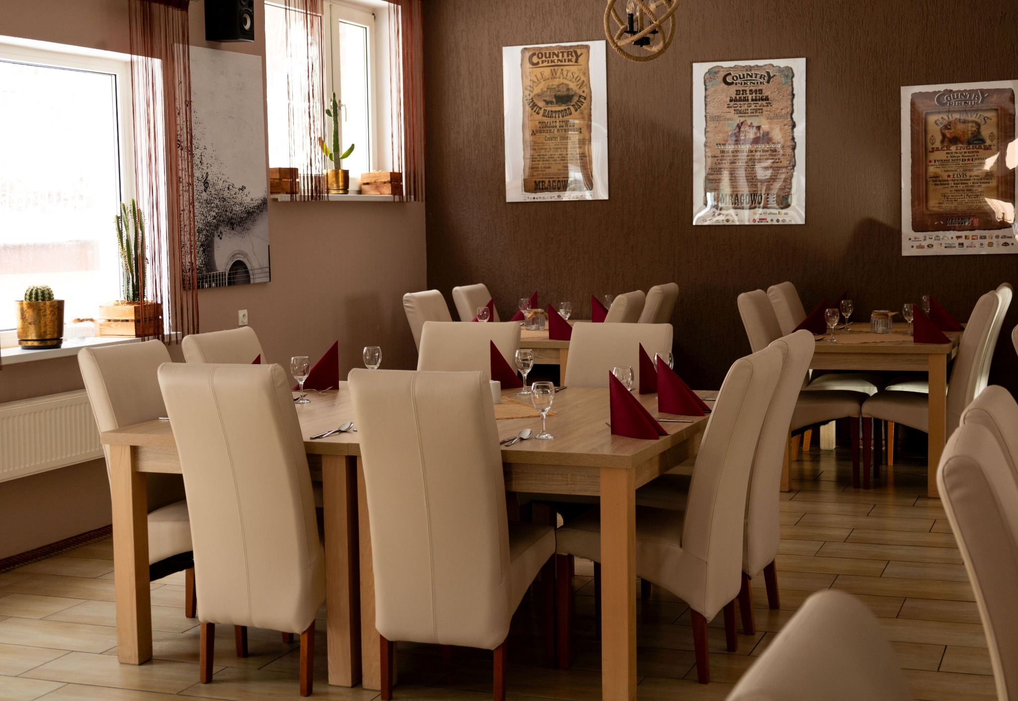 Ustawione stoły z beżowymi krzesłami i czerwonymi serwetkami w lokalu z brązowymi ścianami, na których wiszą plakaty wydarzeń.