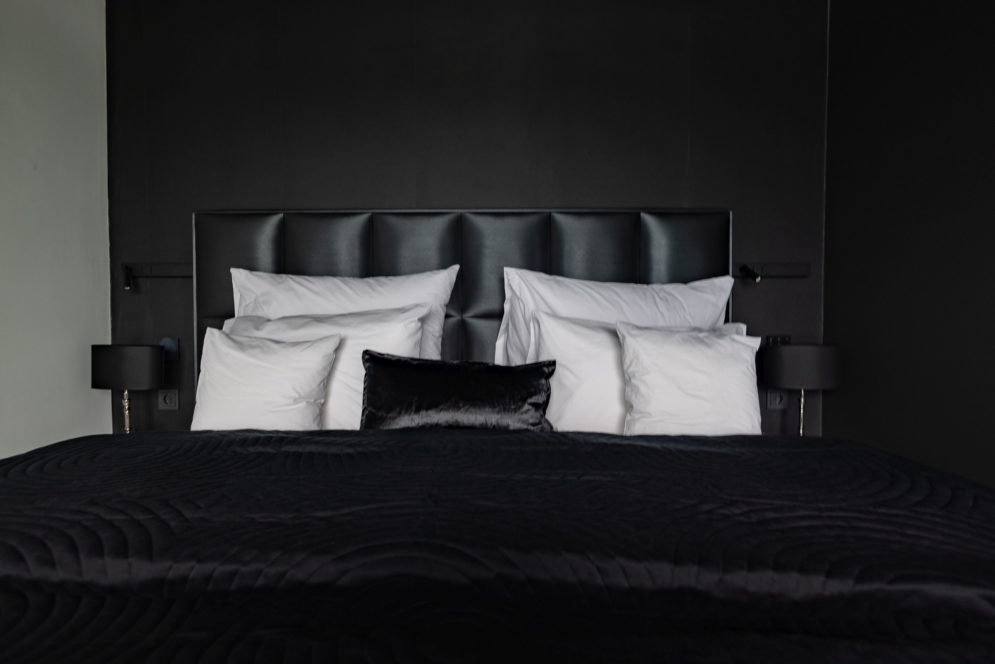 Elegancka sypialnia z czarną tapetą, pikowanym wezgłowiem łóżka i satynową pościelą w odcieniach czerni i bieli, oświetlona lampkami nocnymi z abażurami.