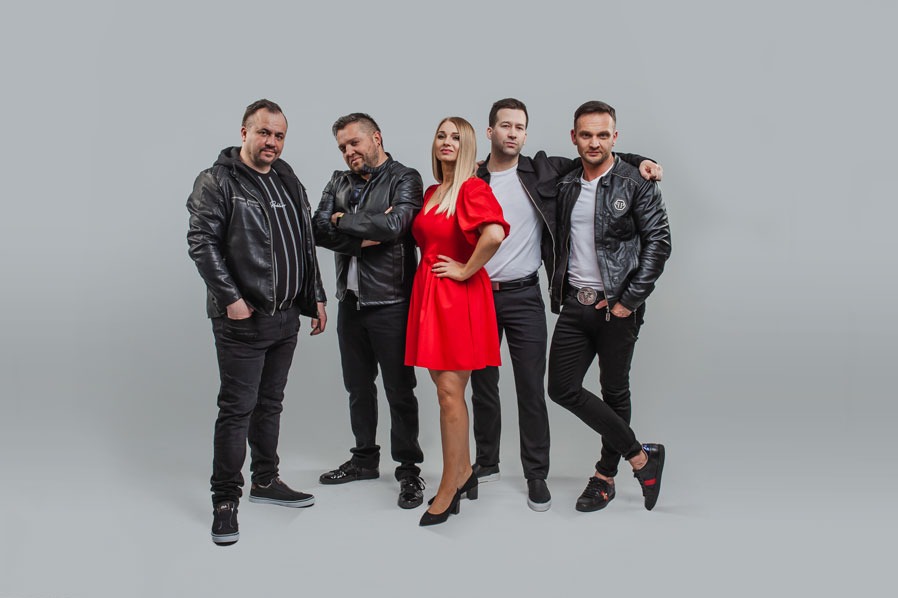 Zespół muzyczny w skórzanych kurtkach i czerwonej sukience pozuje na jasnoszarym tle. Jeden z muzyków ma rękę na ramieniu kolegi. Uwagę przykuwa czerwona sukienka.