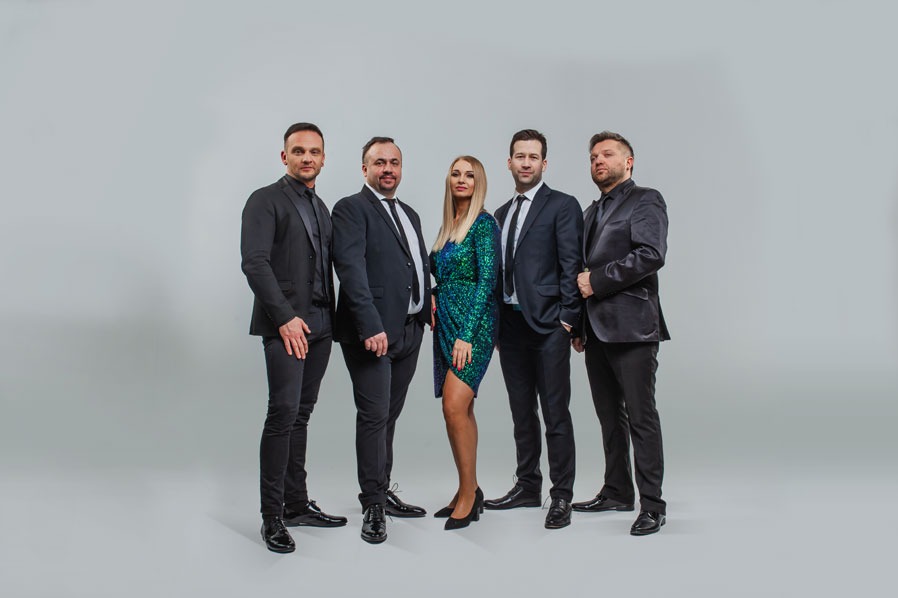 Zespół muzyczny w eleganckich strojach pozuje na jasnym tle. Kobieta w cekinowej sukience pośrodku. Mężczyźni w garniturach z krawatami i bez. Profesjonalna sesja.