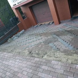 CITY BUD - Ułożona część podjazdu z kostki brukowej w kolorze ceglastym, widoczne niedokończone fragmenty oraz przygotowane kostki do dalszego układania, brama wjazdowa z czarnego metalu po lewej stronie.