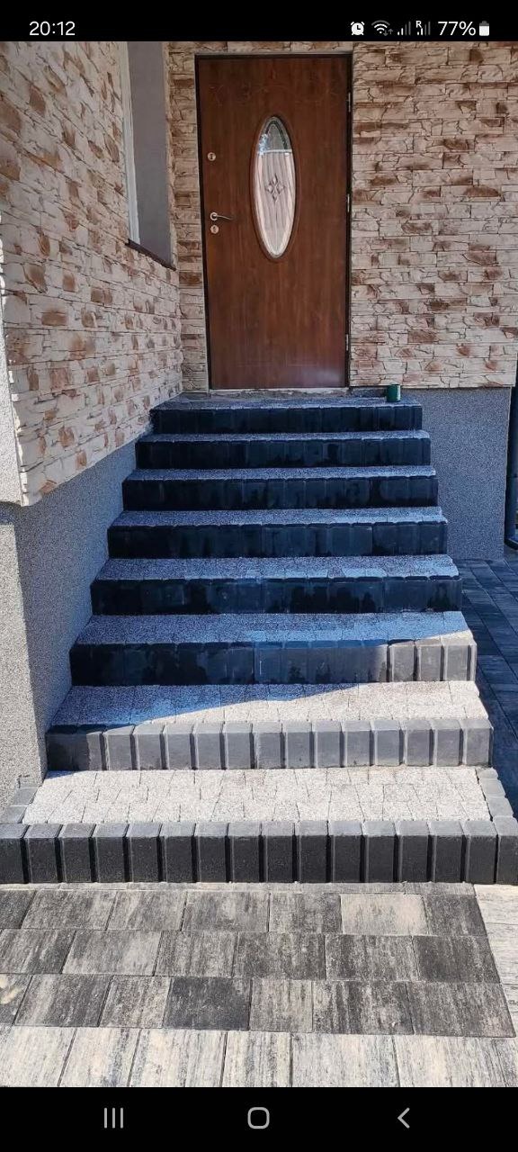Schody wejściowe wyłożone szarą kostką brukową z ciemniejszymi obrzeżami, prowadzące do brązowych drzwi z owalnym oknem, ściana wykończona imitacją kamienia.