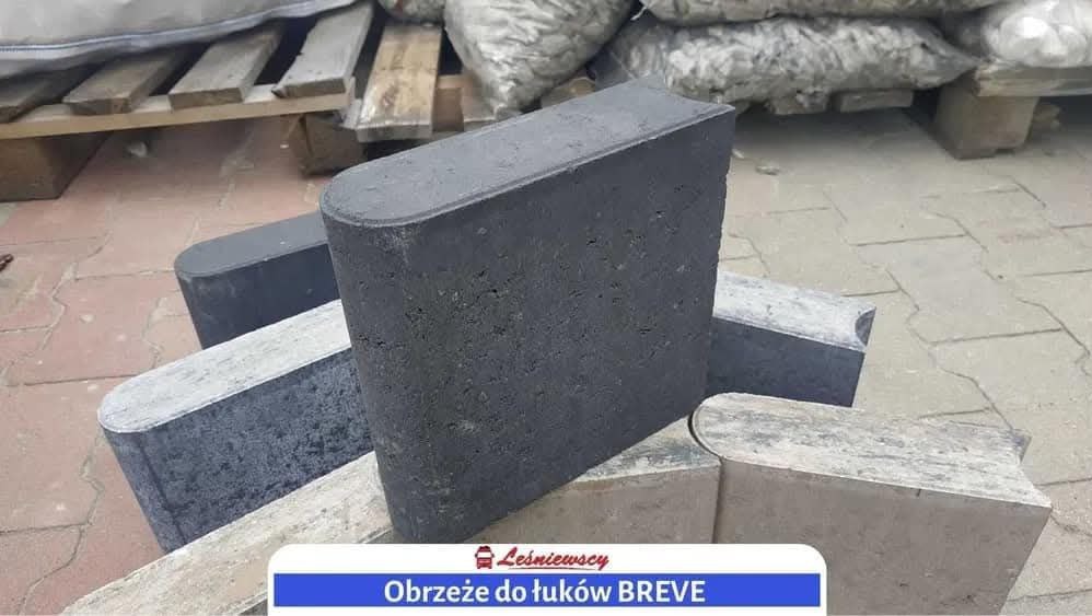 Prezentacja obrzeży betonowych BREVE w odcieniach szarości, ułożonych na kostce brukowej, z paletą i workami z kamieniem w tle.