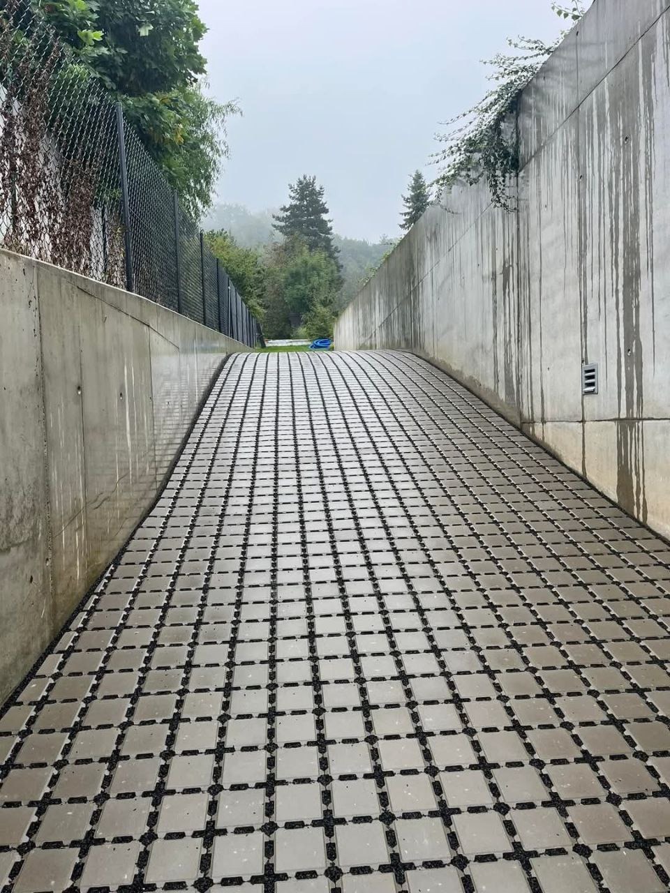 Ułożona kostka na rampie w betonowym tunelu, widok w perspektywie z lekkim nachyleniem, drzewa w tle.