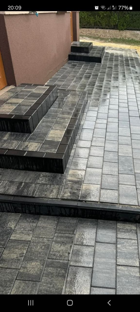 Ułożone schody z kostki brukowej w odcieniach szarości, z czarnymi obrzeżami, prowadzące do budynku o brązowej elewacji.