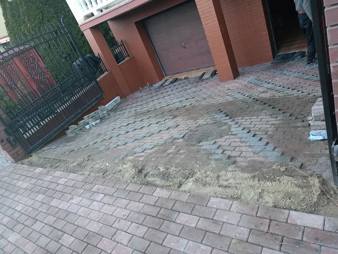 Ułożona część podjazdu z kostki brukowej w kolorze ceglastym, widoczne niedokończone fragmenty oraz przygotowane kostki do dalszego układania, brama wjazdowa z czarnego metalu po lewej stronie.