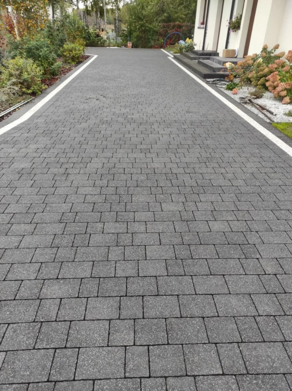 Szeroki podjazd z szarej kostki brukowej ułożonej we wzór jodełki, z białymi obrzeżami, prowadzący do domu z jasną elewacją i zielenią w tle.