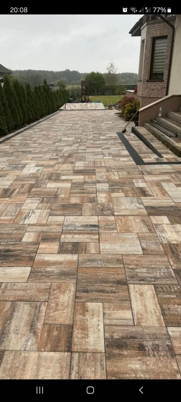 Ułożona kostka brukowa w odcieniach brązu i beżu, tworząca wzór prostokątów na podjeździe przed domem z zielonym żywopłotem w tle.