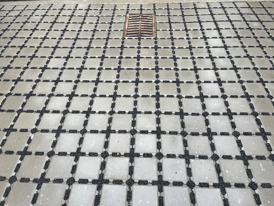 Ułożona nawierzchnia z betonowych kwadratów z czarną, drobną frakcją wypełniającą przestrzenie między nimi, z centralnie umieszczoną kratką ściekową w górnej części kadru.