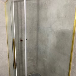 SERHII - BUD SERHII PINKO - Kabina prysznicowa z przesuwnymi drzwiami i złotymi profilami na tle szarych płytek w łazience. Widok z boku na detale wykończenia.