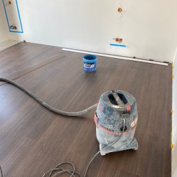 SERHII - BUD SERHII PINKO - Układanie paneli: ciemne panele podłogowe, odkurzacz przemysłowy i listwy przypodłogowe przy ścianie. Widoczne gniazdka elektryczne zabezpieczone taśmą.