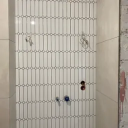 Ściana w łazience z białymi, podłużnymi płytkami ułożonymi pionowo. Widoczne otwory na armaturę i surowe wykończenie ściany obok.