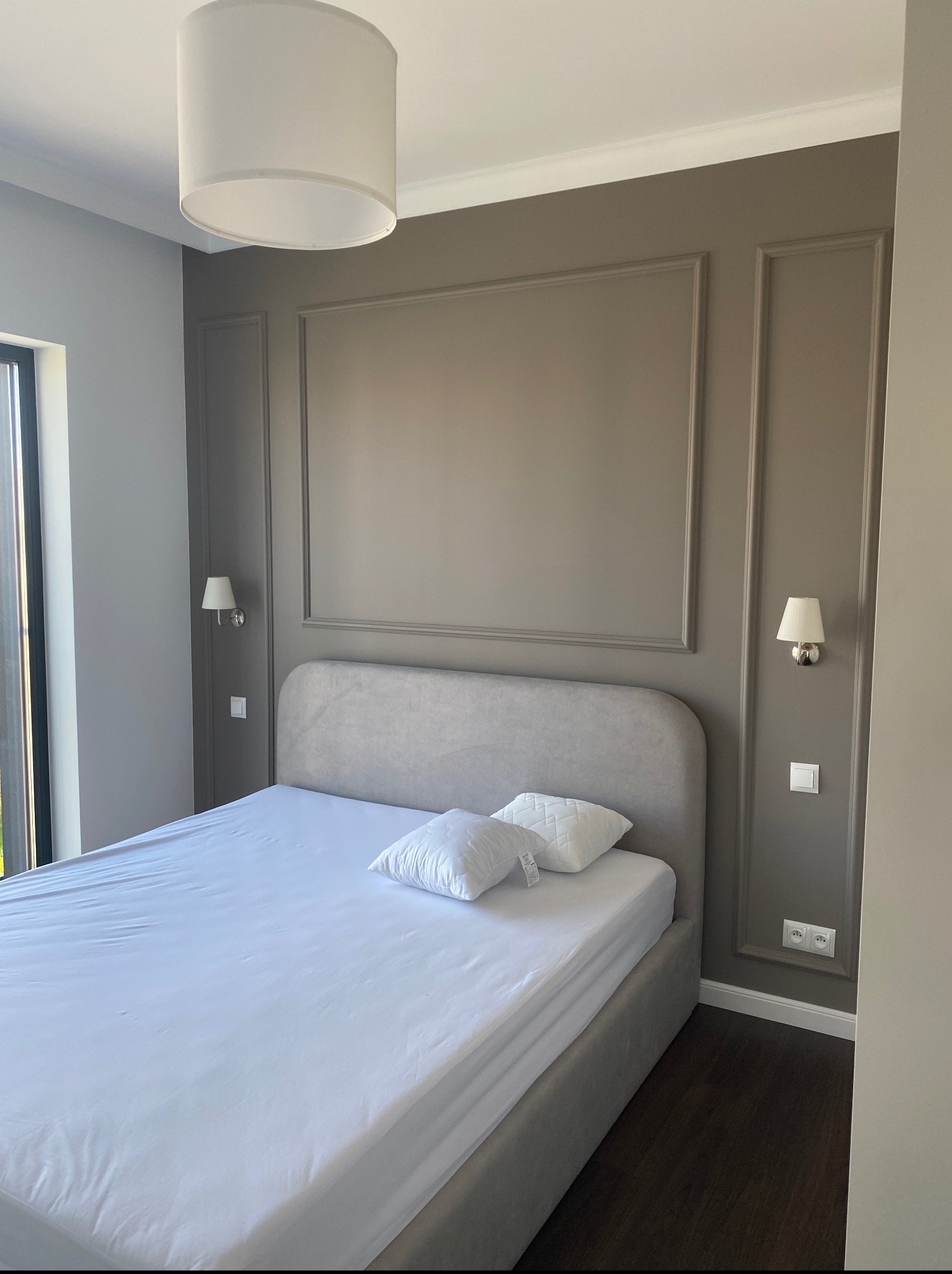 Elegancka sypialnia w stonowanych szarościach i brązach. Dekoracyjne listwy na ścianach, minimalistyczne lampy ścienne i sufitowa. Podłoga z ciemnego drewna.