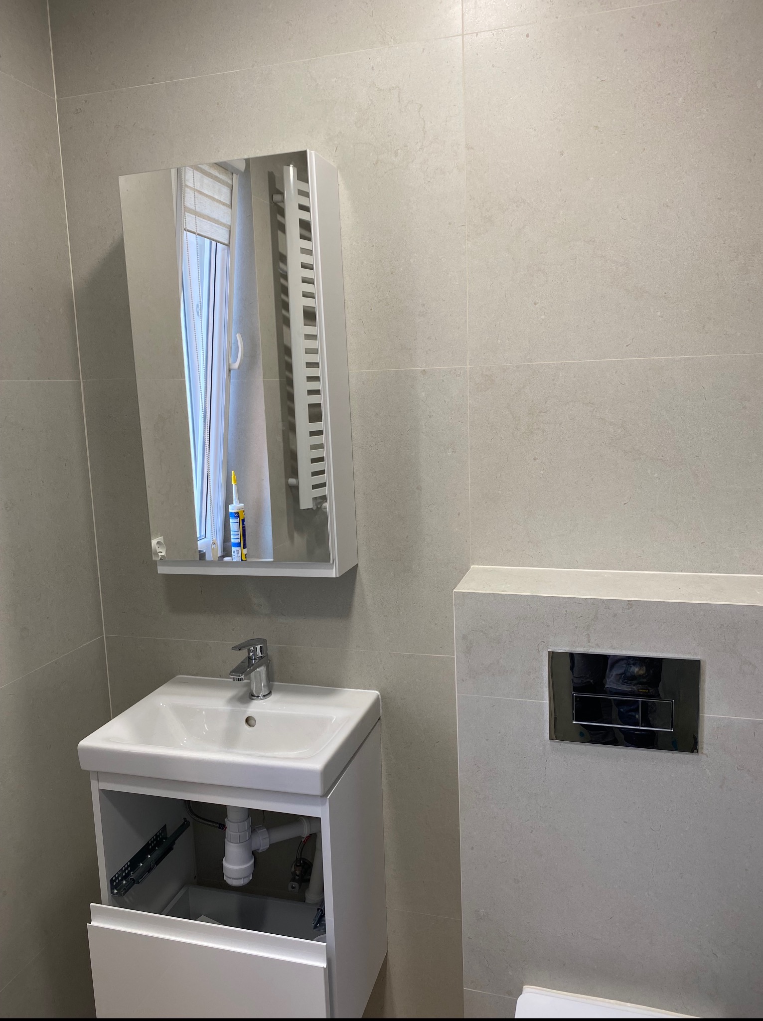 Minimalistyczna łazienka z umywalką, lustrem i płytkami w odcieniach szarości. Częściowo otwarta szafka pod umywalką ukazuje elementy instalacji.