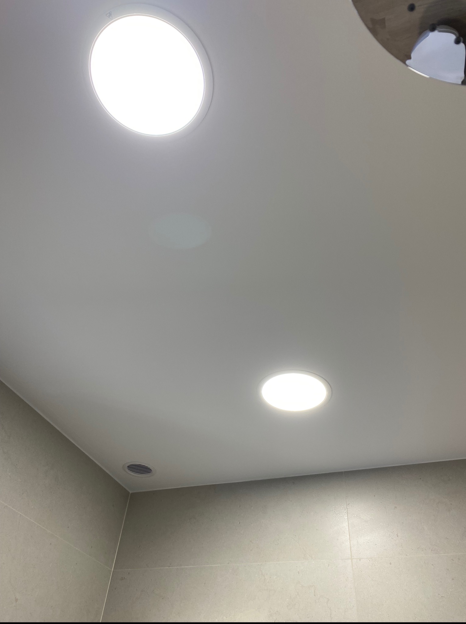 Minimalistyczna łazienka: gładki, szary sufit z dwoma okrągłymi lampami LED i kratką wentylacyjną, szare płytki na ścianach. Czyste linie i nowoczesny design.