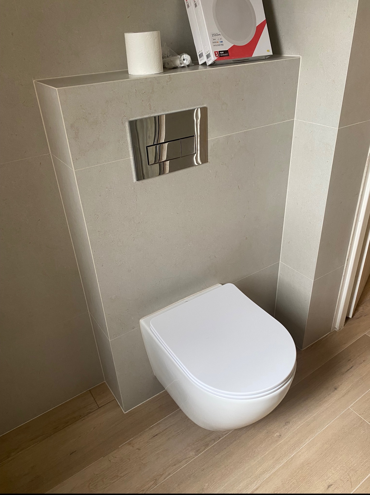 Wisząca toaleta z szarą zabudową, chromowanym przyciskiem i rolką papieru toaletowego na górze. Podłoga wyłożona panelami imitującymi drewno.