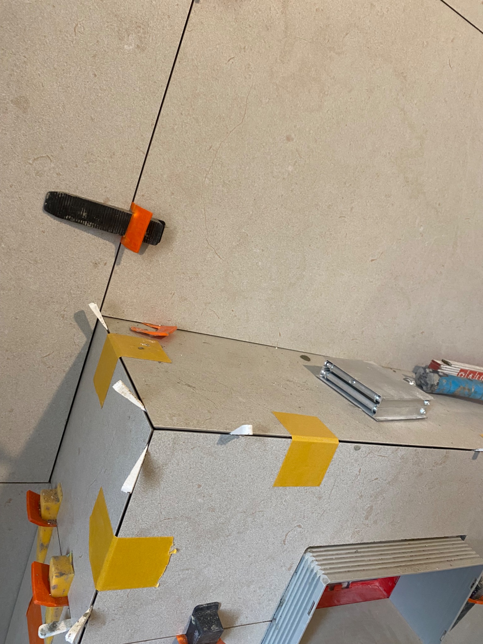 Świeżo ułożone, beżowe płytki z systemem poziomowania i żółtą taśmą. Widoczne narzędzia glazurnicze i fragment instalacji. Detal wykończenia.