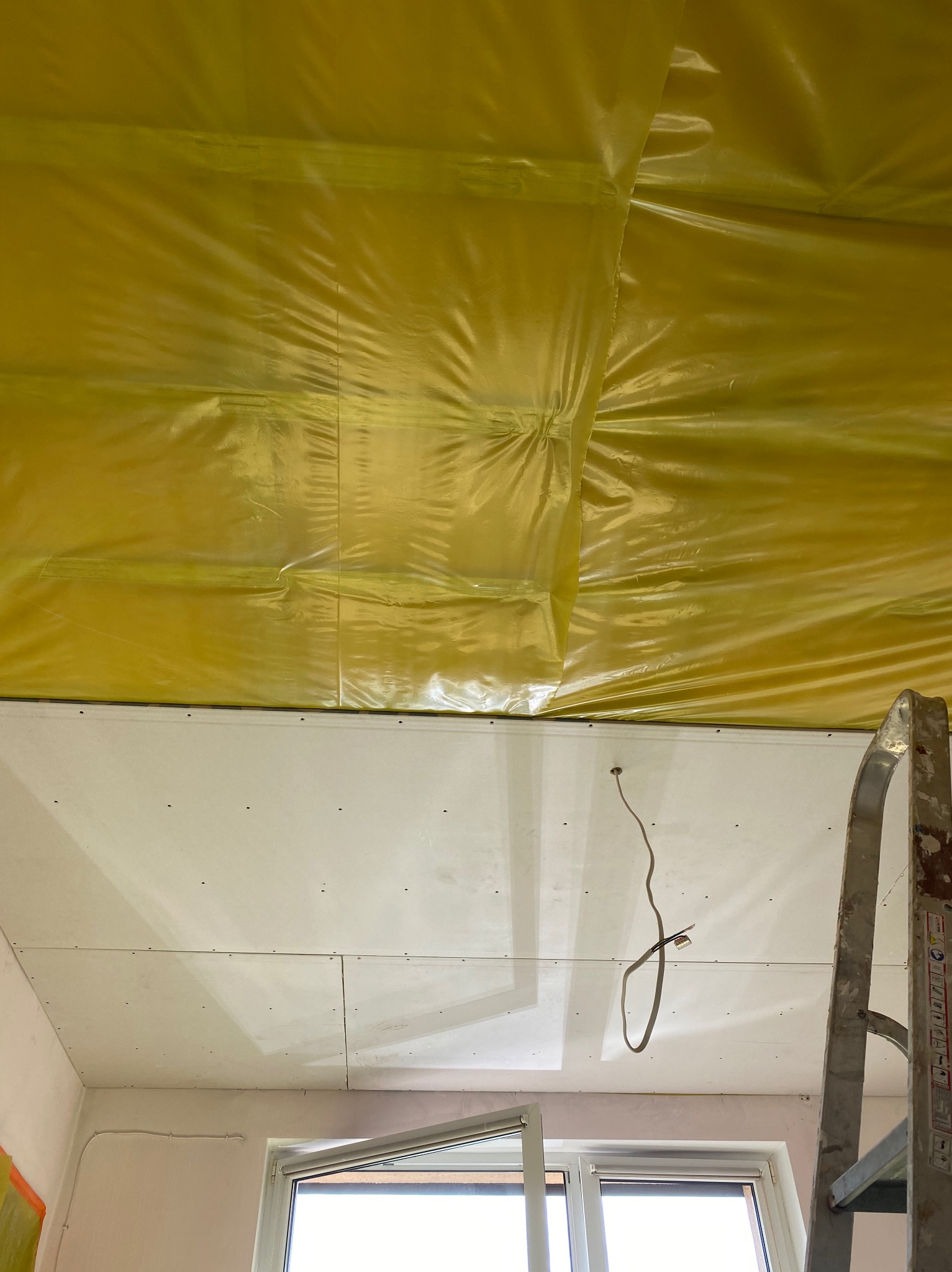 Żółta folia izolacyjna i płyty gipsowe na suficie, widoczne przewody elektryczne i fragment drabiny. Etap prac wykończeniowych w pomieszczeniu.
