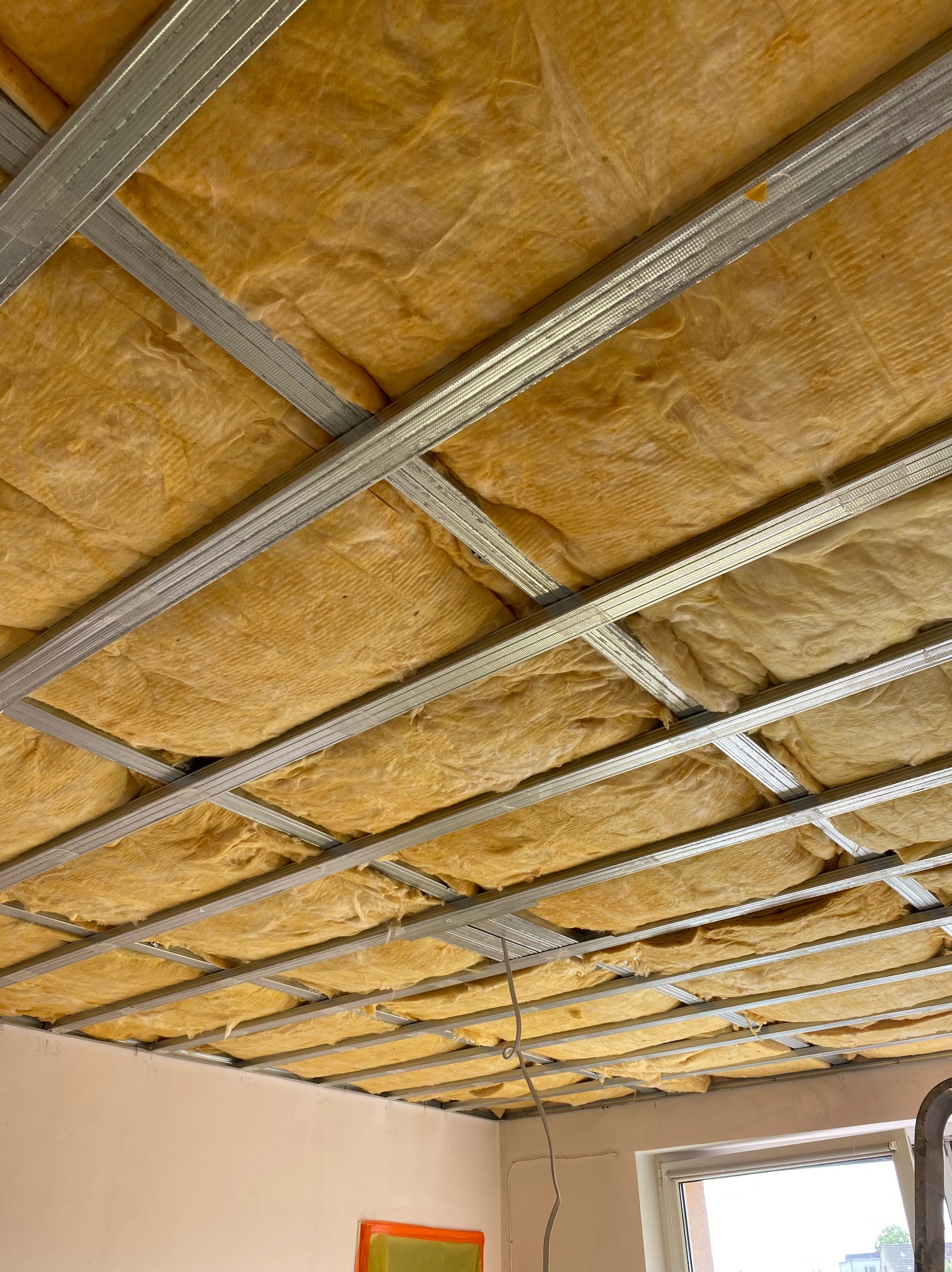 Montaż izolacji z wełny mineralnej w metalowym stelażu sufitu podwieszanego. Widoczne kable i fragment drabiny. Ocieplenie stropu w trakcie prac.