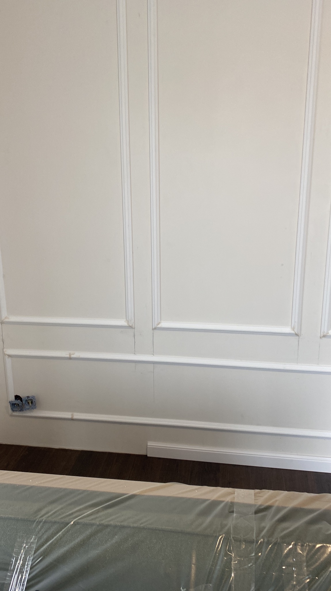 Ściana z dekoracyjnymi panelami ściennymi w stylu klasycznym, z widocznymi puszkami elektrycznymi i podłogą. Częściowo widoczny materiał budowlany pod folią.