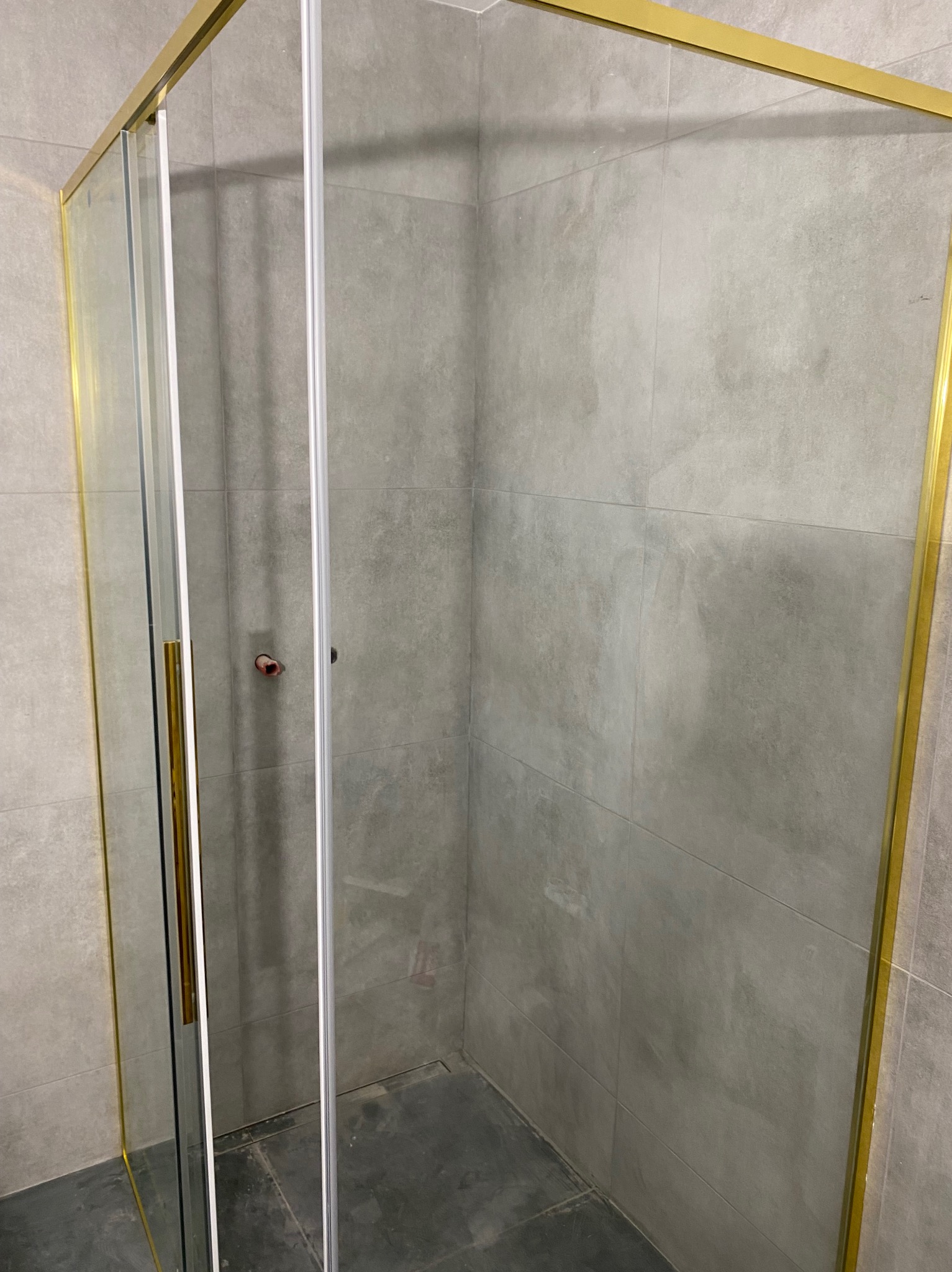 Kabina prysznicowa z przesuwnymi drzwiami i złotymi profilami na tle szarych płytek w łazience. Widok z boku na detale wykończenia.