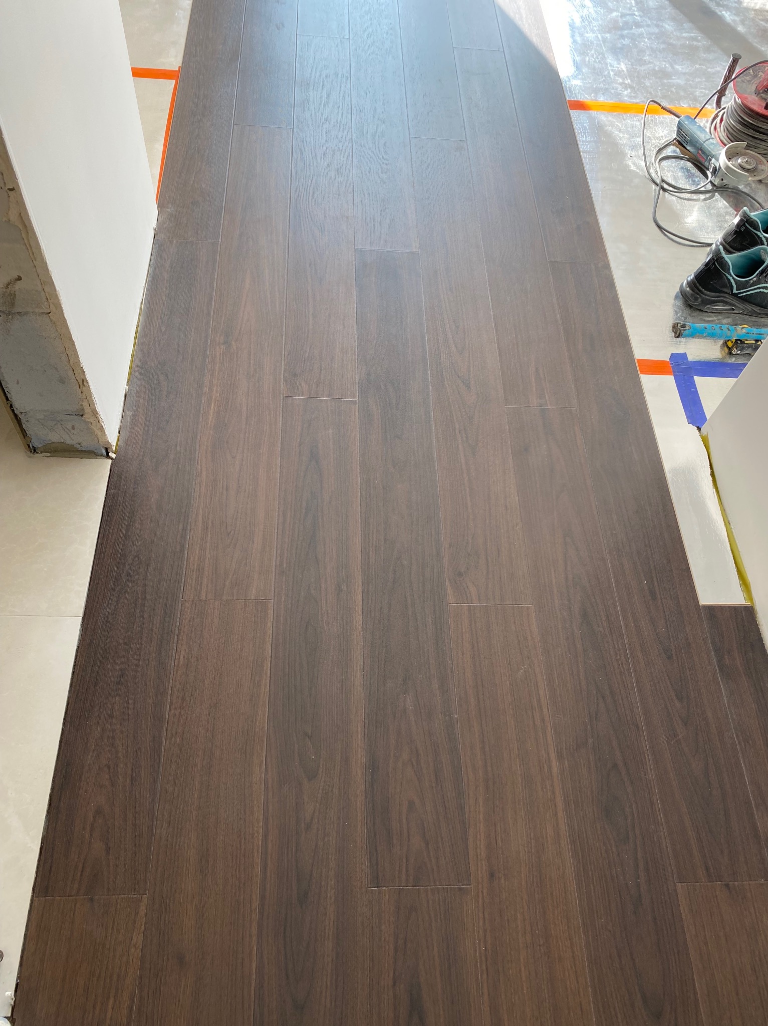 Ułożone, ciemne panele podłogowe w trakcie montażu, widoczne narzędzia i buty robocze na podłożu zabezpieczonym folią. Prace wykończeniowe, widok z góry.