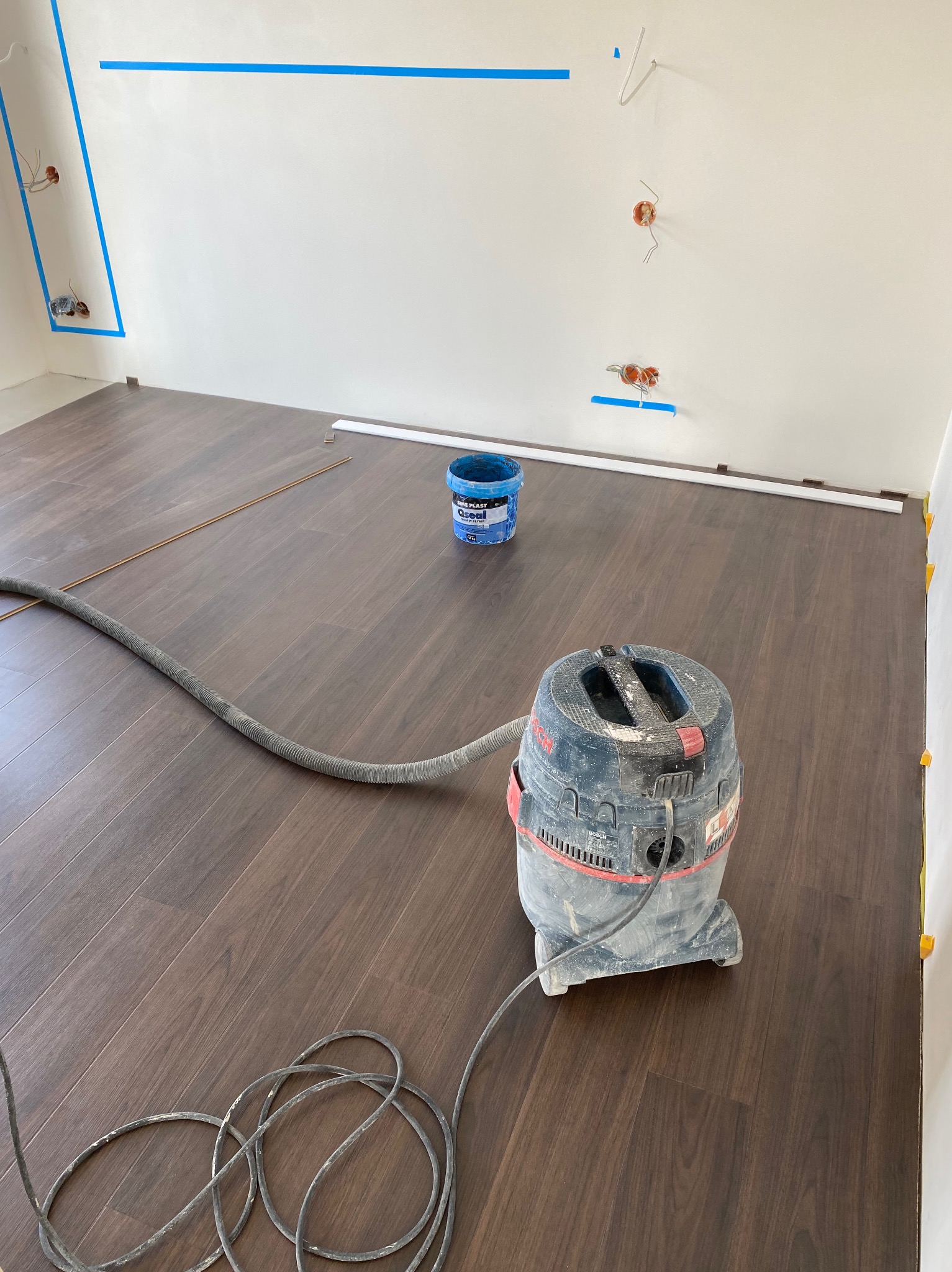 Układanie paneli: ciemne panele podłogowe, odkurzacz przemysłowy i listwy przypodłogowe przy ścianie. Widoczne gniazdka elektryczne zabezpieczone taśmą.