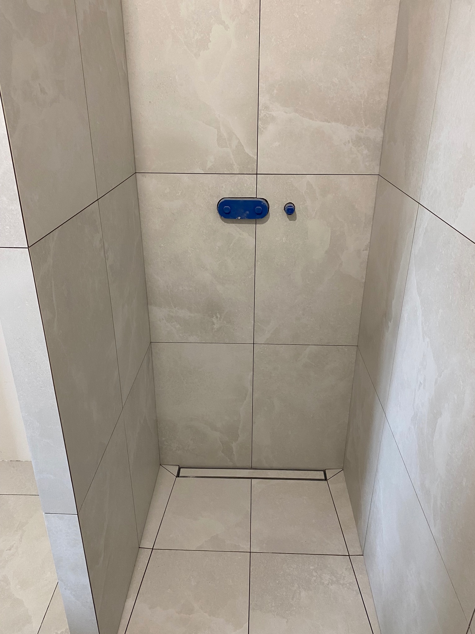 Wykończona kabina prysznicowa z beżowymi płytkami imitującymi marmur, z odpływem liniowym i niebieskim przyciskiem do spłukiwania toalety podtynkowej.