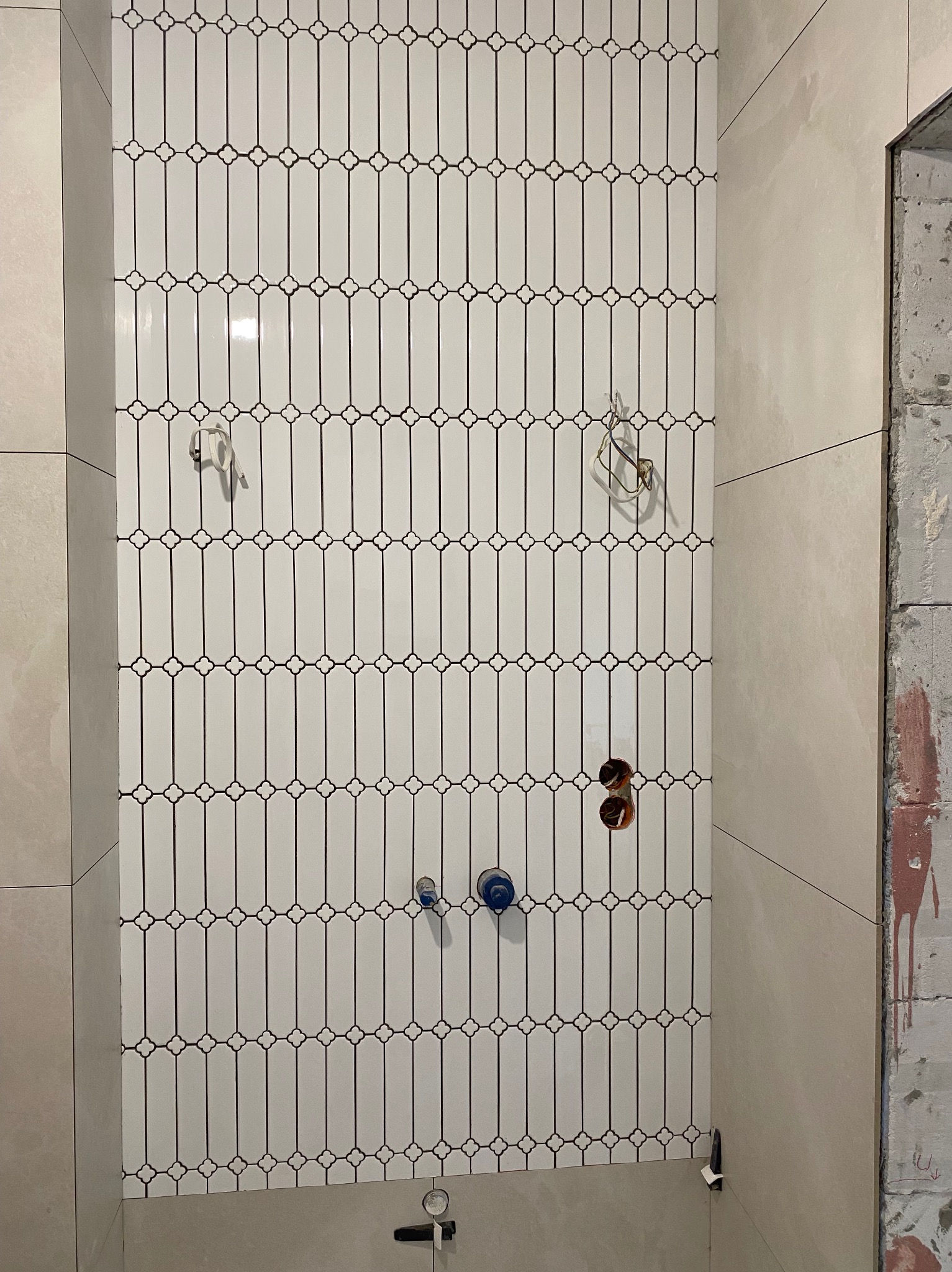 Ściana w łazience z białymi, podłużnymi płytkami ułożonymi pionowo. Widoczne otwory na armaturę i surowe wykończenie ściany obok.