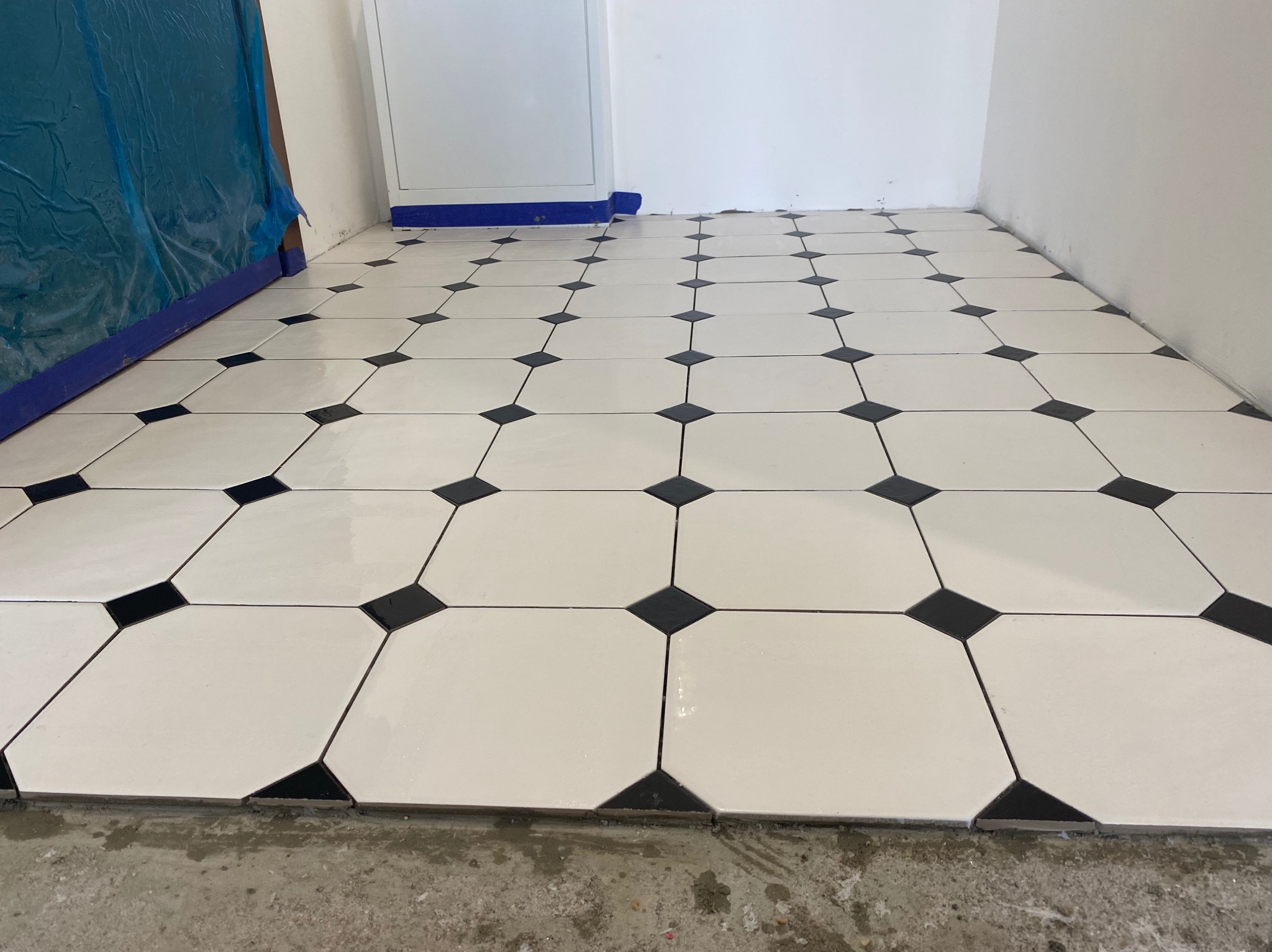 Biało-czarna podłoga z płytek ułożonych w romby, widok z góry, świeżo po ułożeniu, z widocznymi zabezpieczeniami przy ścianie i fragmentem surowej podłogi.
