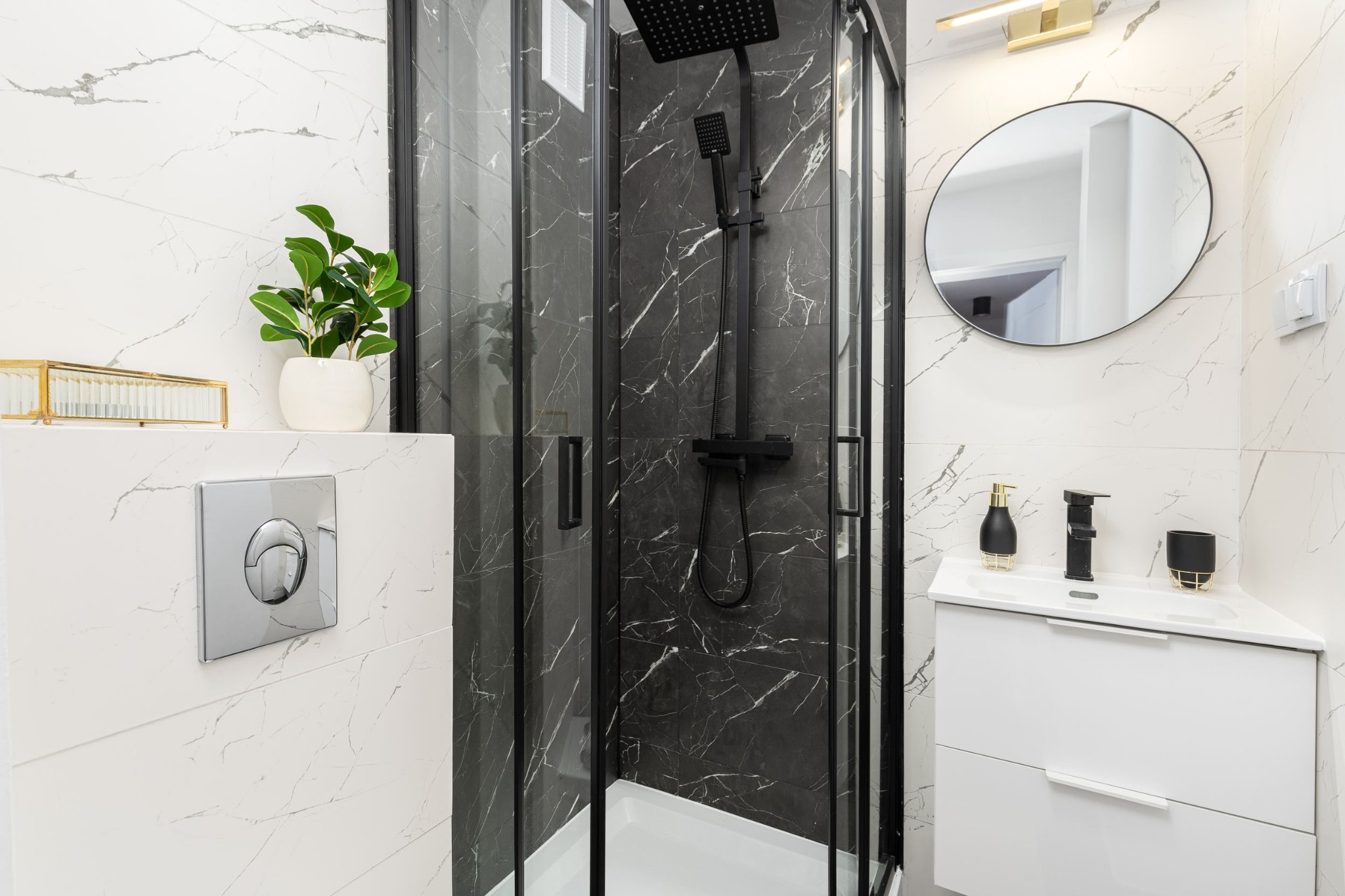 Nowoczesna łazienka z czarno-białymi marmurkowymi płytkami, kabiną prysznicową typu walk-in z czarną armaturą, białą szafką umywalkową i okrągłym lustrem z czarną ramą.