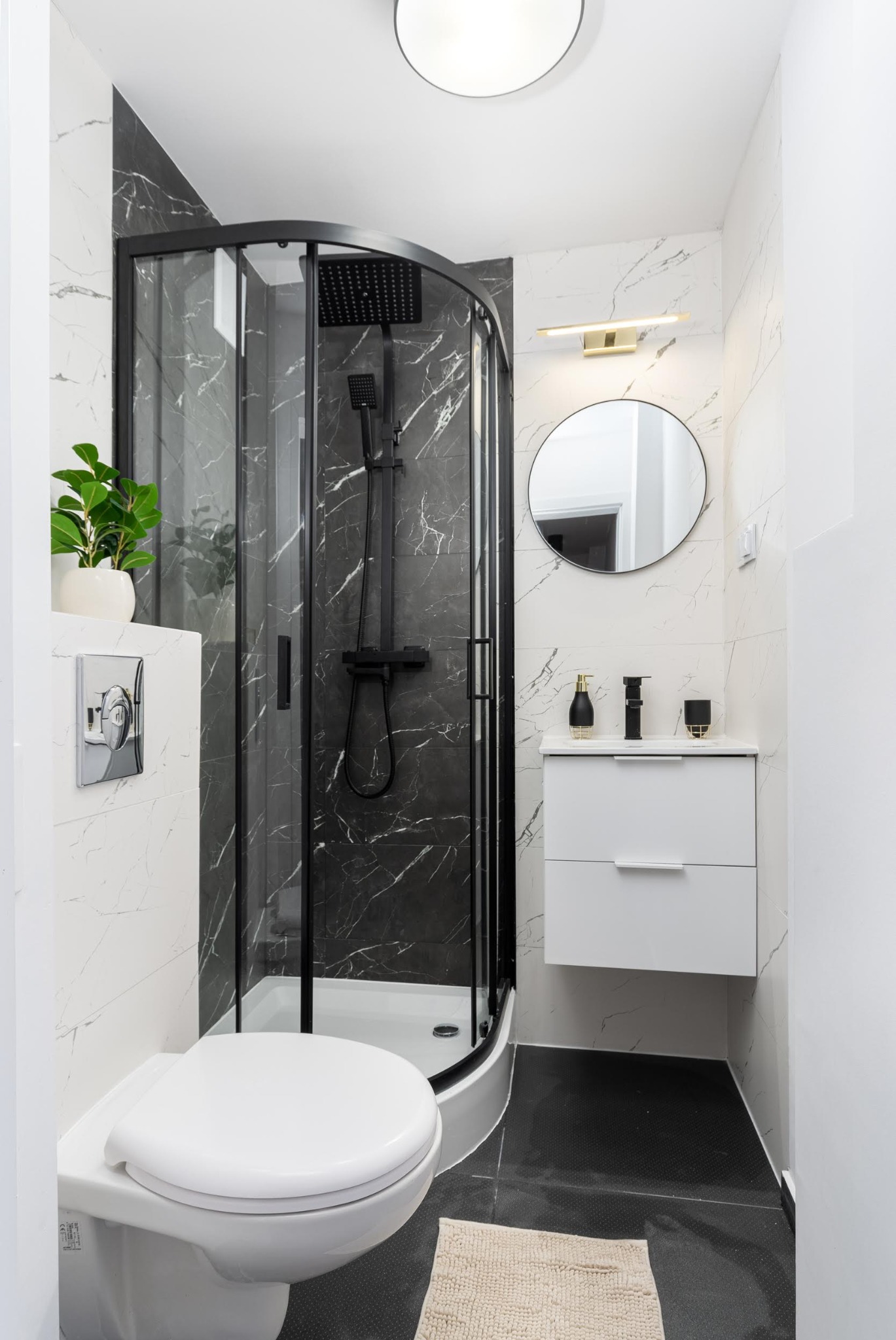 Nowoczesna łazienka z kabiną prysznicową w czarnej ramie, marmurowe płytki na ścianach, czarna podłoga, wisząca szafka z umywalką i okrągłym lustrem, oraz toaleta z podtynkowym systemem spłukiwania.