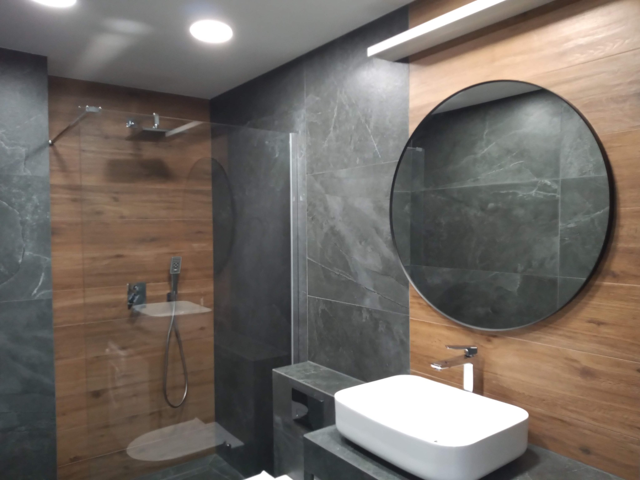 Nowoczesna łazienka z kabiną prysznicową typu walk-in, okrągłym lustrem i umywalką nablatową, wykończona płytkami imitującymi drewno i szarym kamieniem.