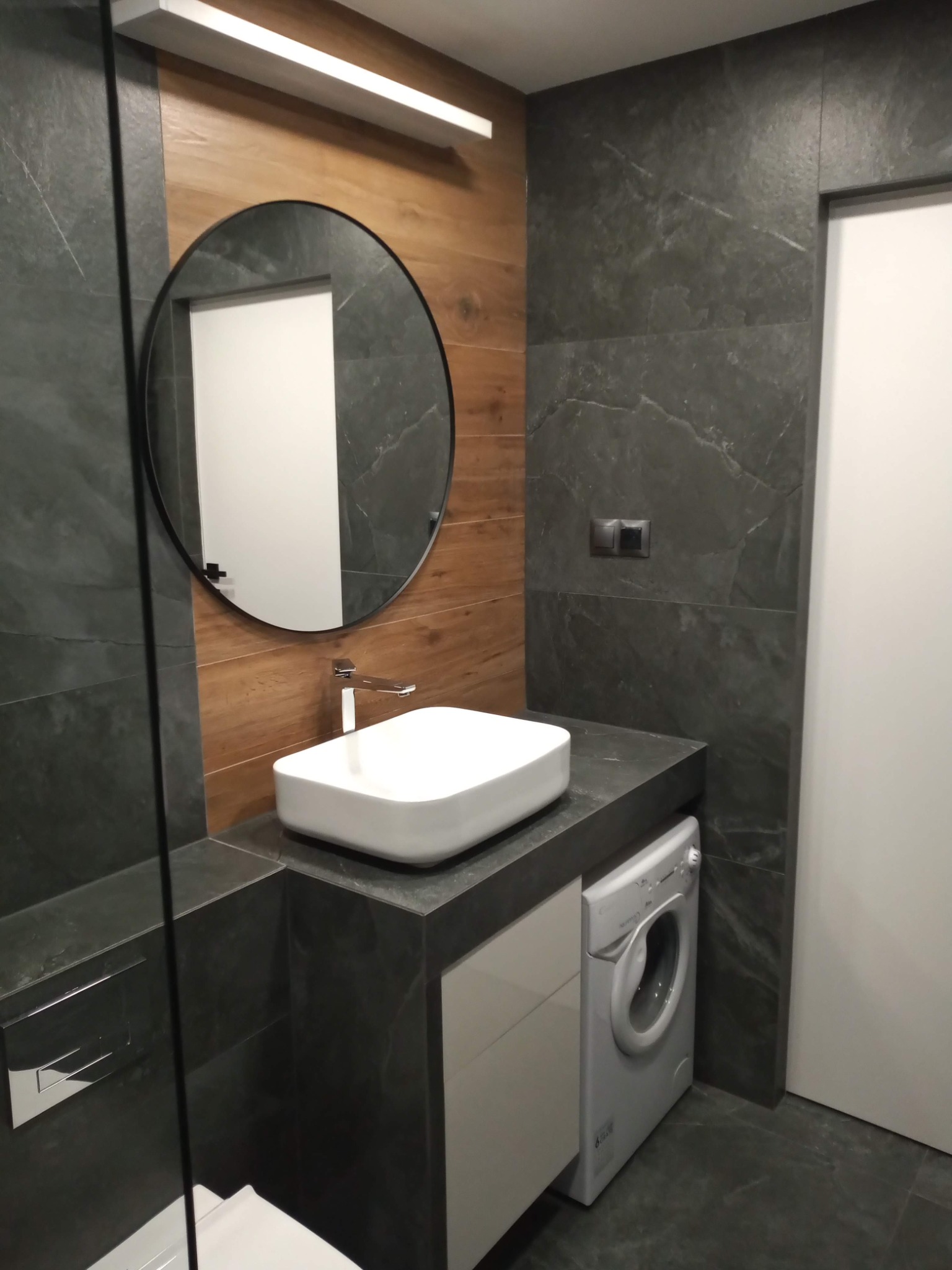 Nowoczesna łazienka z umywalką nablatową, okrągłym lustrem z czarną ramą, szarą płytką imitującą kamień, drewnianą ścianą i pralką w zabudowie.