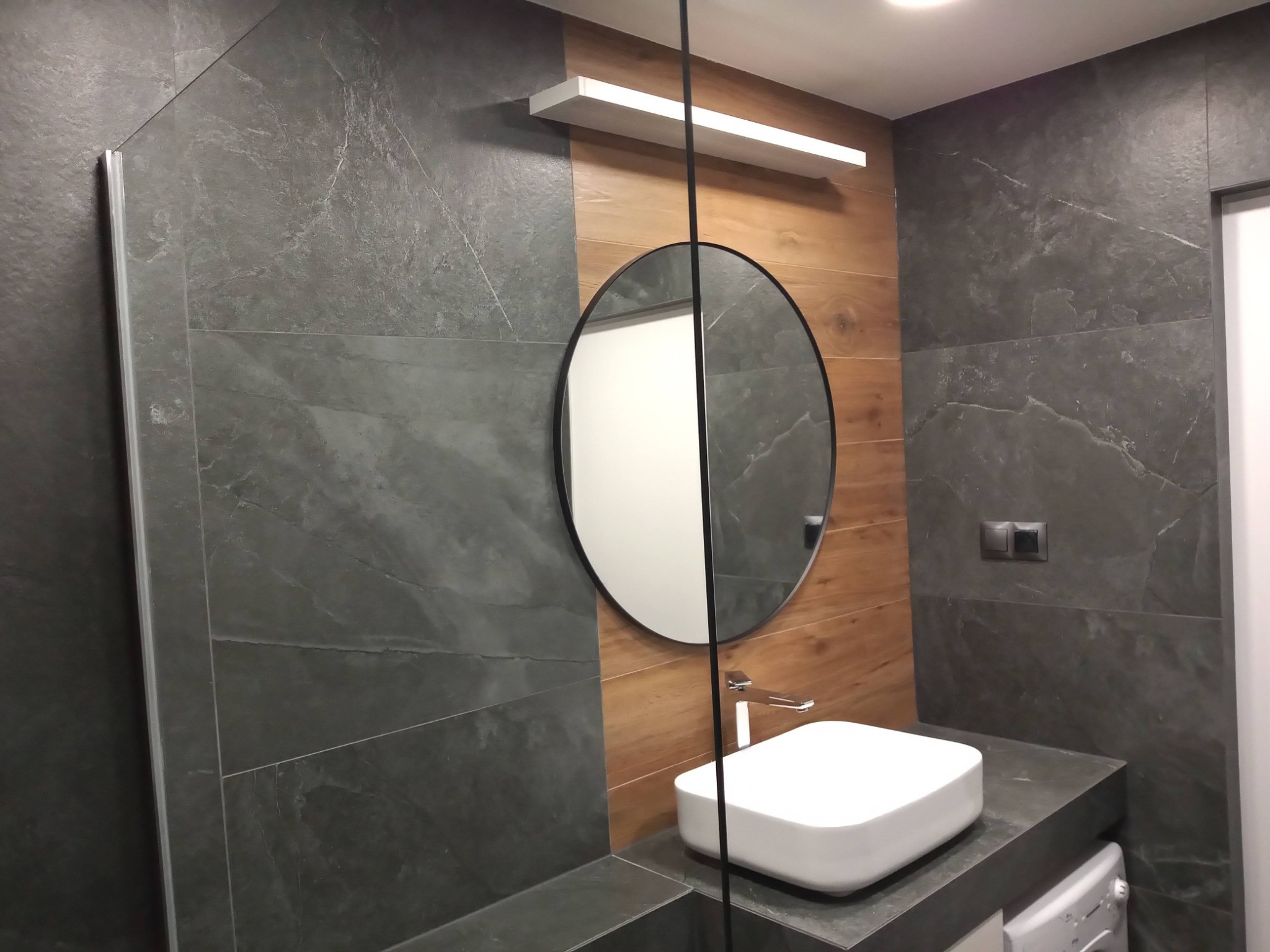 Nowoczesna łazienka z szarymi płytkami imitującymi kamień, okrągłym lustrem i drewnianym akcentem na ścianie, biała umywalka nablatowa i chromowana bateria, widoczna część kabiny prysznicowej.